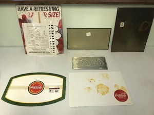 lot 101 image: 2 Coca Cola metal signs, 1 license plate, 1 cardboard standup sign still in plastic and 2 vinyl placemats