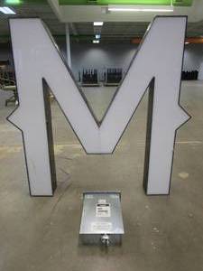 lot 100 image: Lighted Letter M Outdoor Signage With Transformer. Approximate Dimensions 44 x 5.25 x 42.5 Tall - USED FOR RETAIL BRAND MN HOME OUTLET.