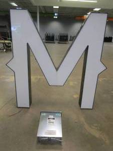 lot 112 image: Lighted Letter M Outdoor Signage With Transformer. Approximate Dimensions 44 x 5.25 x 42.5 Tall - USED FOR RETAIL BRAND MN HOME OUTLET.