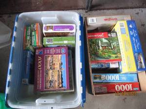 lot 57 image: (1) Fruit box and (1) clamshell closing Storage Tote filled with a nice assortment of jigsaw puzzles in a variety of difficulty levels. (If any pieced were to be missing, it seems to be noted)