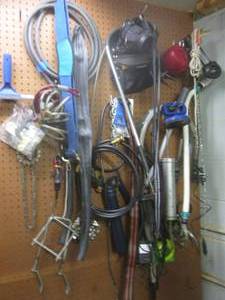 Content on wall consisting of chainsaw blade, bungee cords, grease gun, clamps, cables, fish nets, propane adapter, gardening tools and more