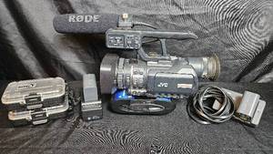 lot 115 image: JVC GY-HM100U Handheld Camcorder with Many Accessories. Rode shotgun microphone, extra batteries with charger, SD card carry cases, and much more.