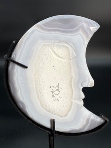 lot 11 image: A luminous Botswana Agate druzy carved into a moon shape, showcasing delicate bands of soft gray, caramel, and cream. The sparkling druzy surface catches the light, in a custom metal stand. 6.6 oz.  0.188 kg.