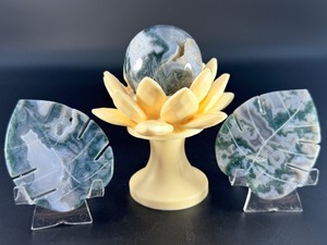 lot 24 image: A serene moss agate collection featuring two hand-carved leaf-shaped pieces and a polished sphere with a druzy center resting on a lotus stand. 11.7 oz.  0.334 kg.
