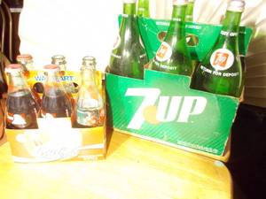 lot 364 image: several bottles of 7UP soda in a vintage-style green cardboard carrier.